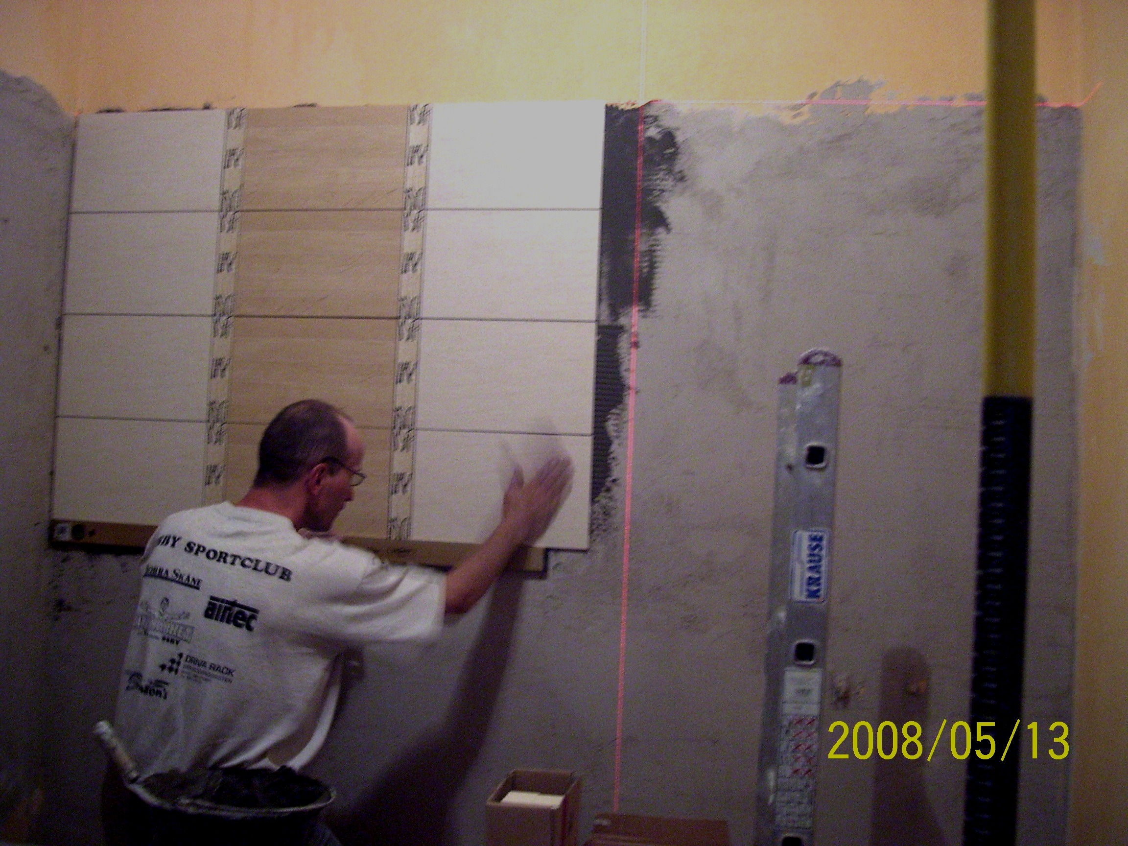 Układanie płytek na ścianie w łazience, widoczny glazurnik dociskający płytkę, pionowy laser wyznaczający linię, drabina Krause w tle, data 2008/05/13.